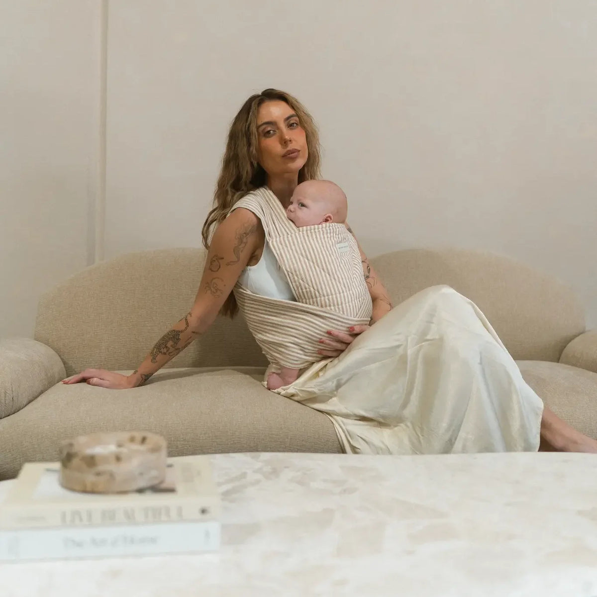 Mum reclining on a cream sofa with her newborn snug in the Burleigh stripe Wrap carrier, one hand under his feet.