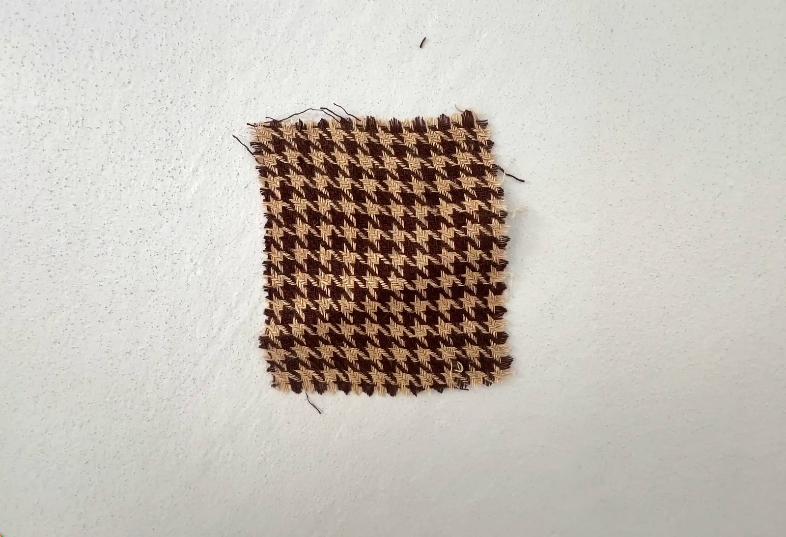 A handwoven tan and chocolate houndstooth linen swatch with raw fringed edges, pinned to a white wall
