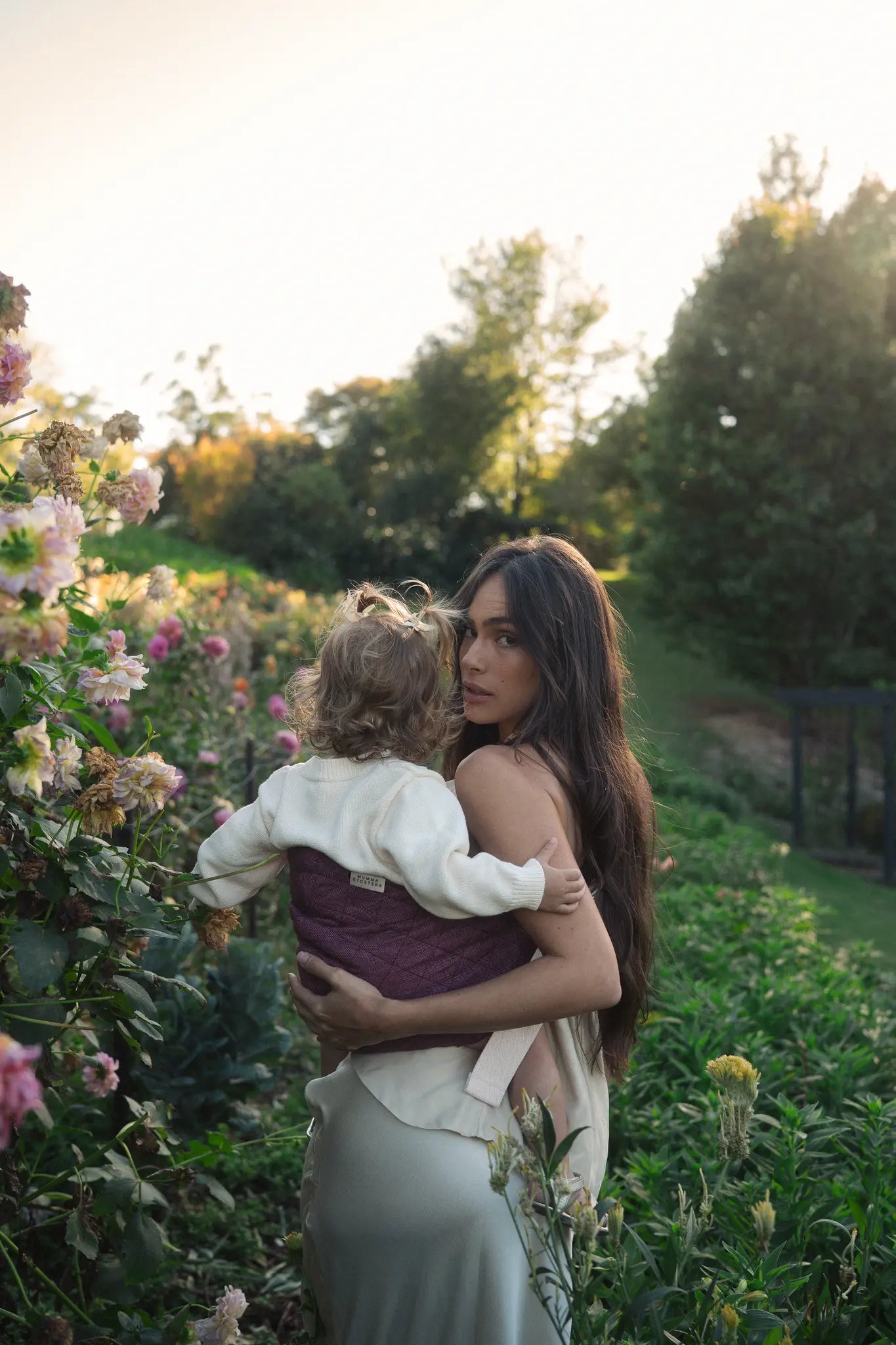 Seen from behind, the Bloomè linen Hipsy toddler carrier cradles a toddler on her mum's hip in a flower field.