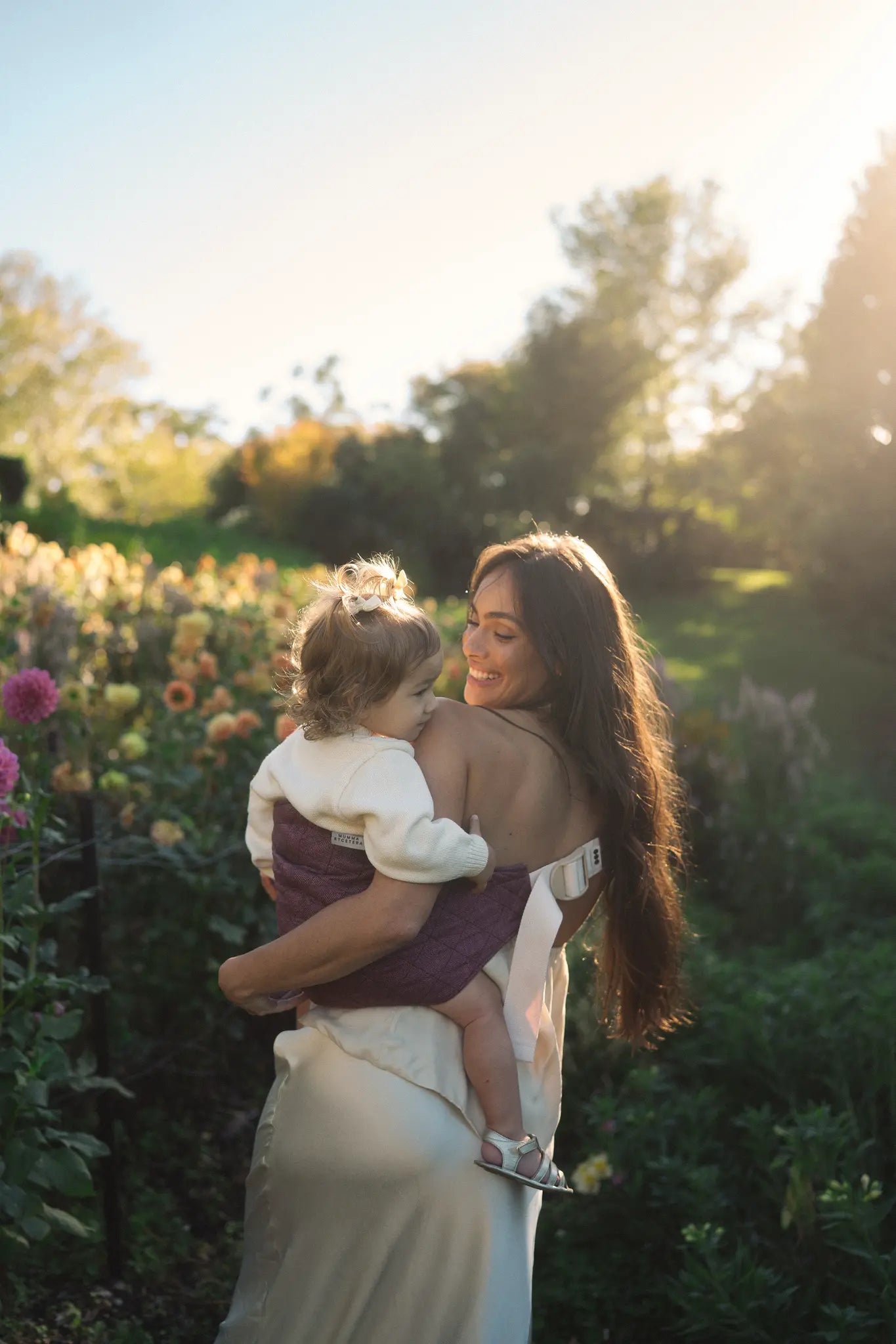 Backlit in the flower farm, a mum steadies her toddler in the Bloomè linen Hipsy toddler carrier.