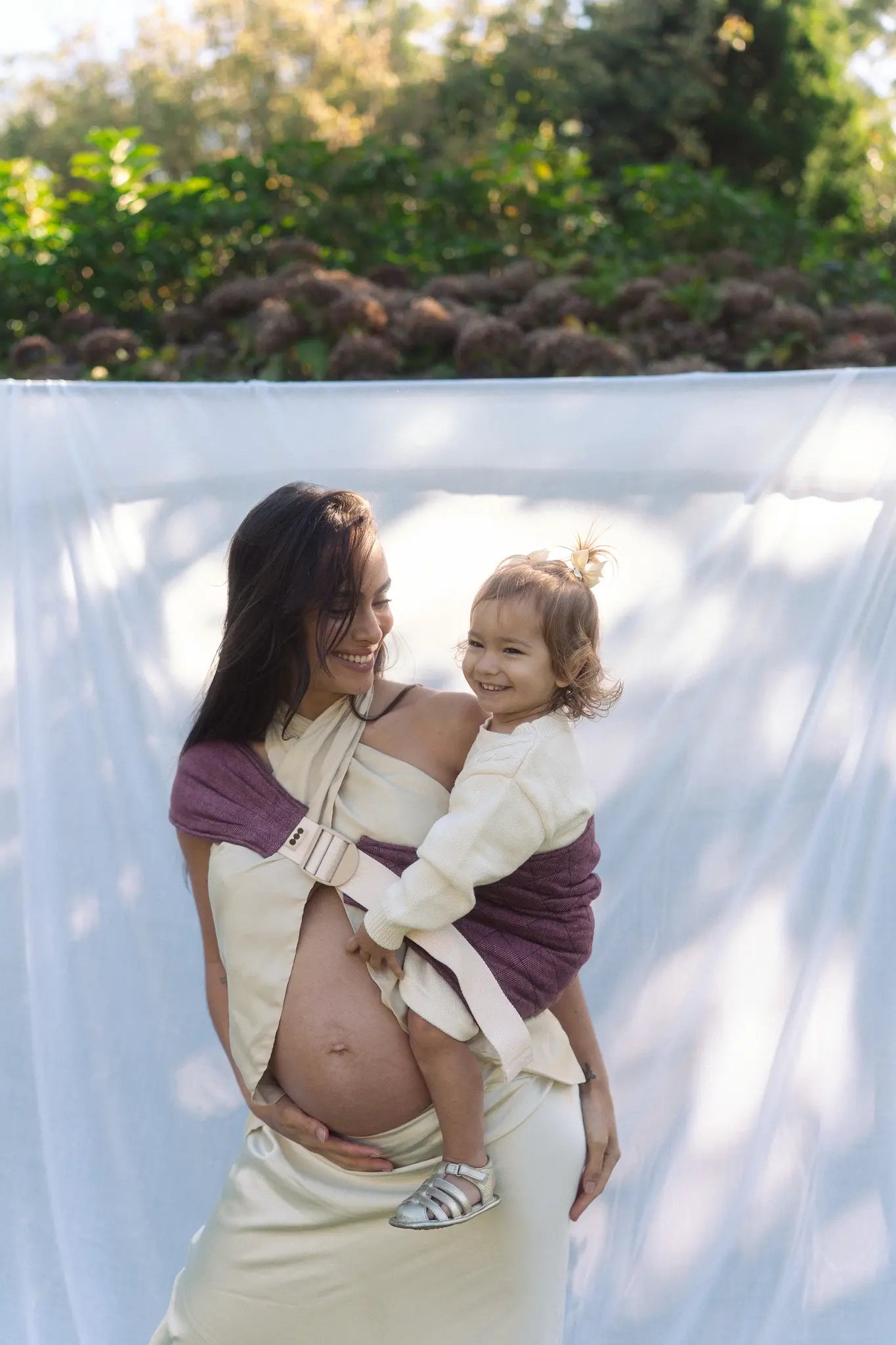 Mum and toddler by a hanging white sheet, her arm around him in the Bloomè linen Hipsy hip carrier