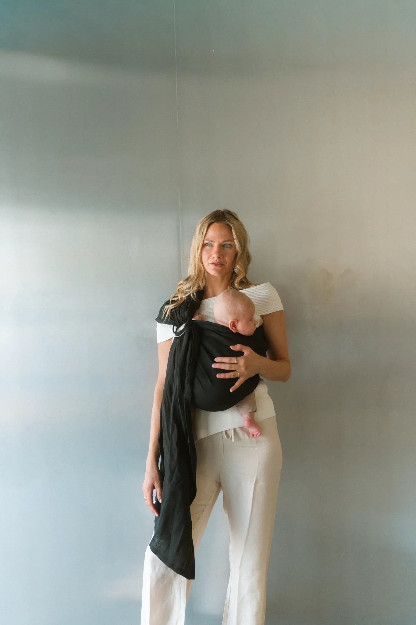 A mum stands against a soft grey wall with her baby held close in the Tao linen Sling baby carrier.