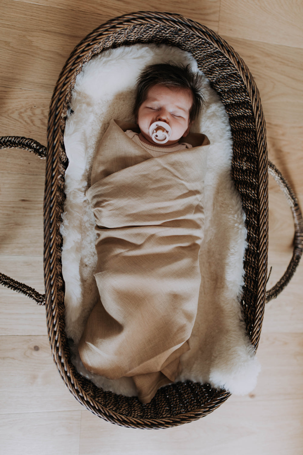 Newborn asleep with a dummy, swaddled in the Bondi linen swaddle on a sheepskin in a cane bassinet