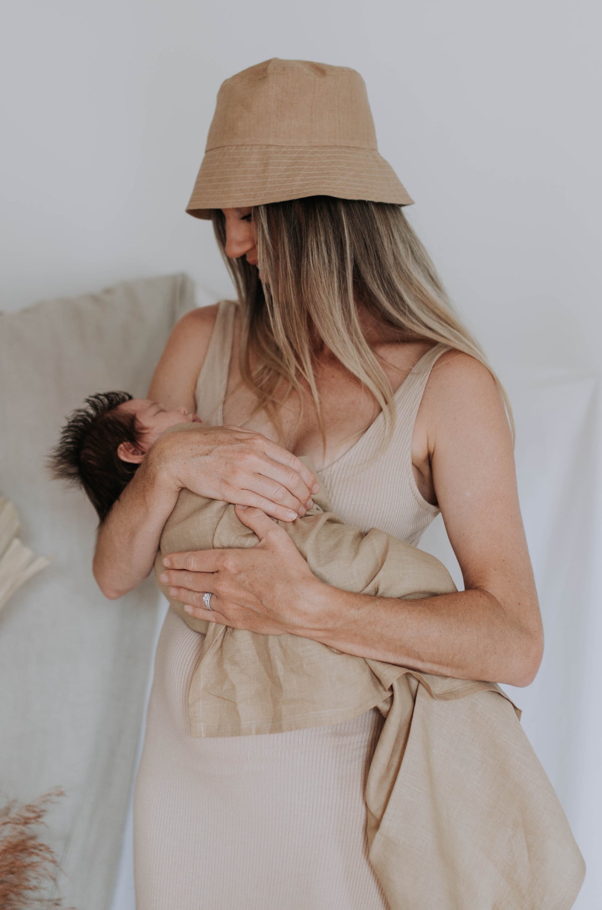 Mum in a sand linen bucket hat cradling her newborn in the Bondi linen swaddle against a linen backdrop