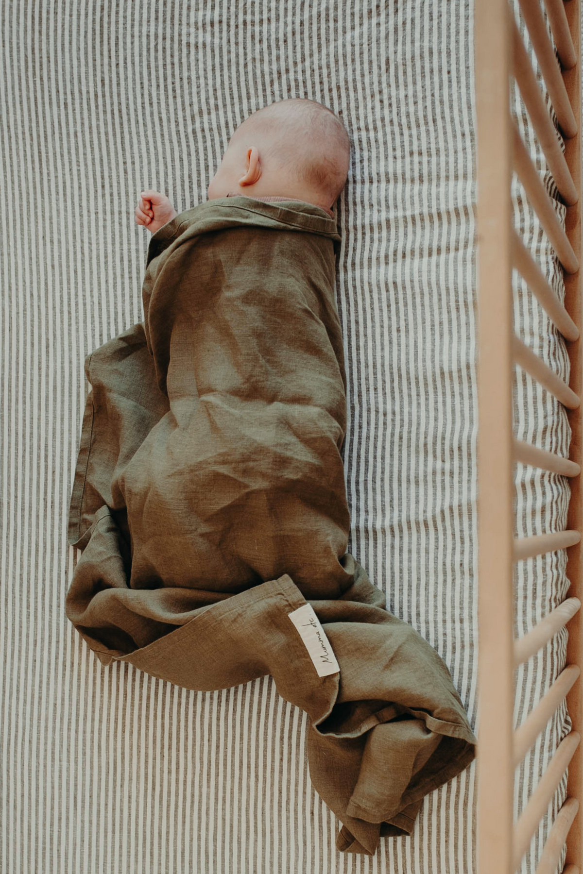Newborn asleep in the Willow linen swaddle, olive green on a striped cot mattress