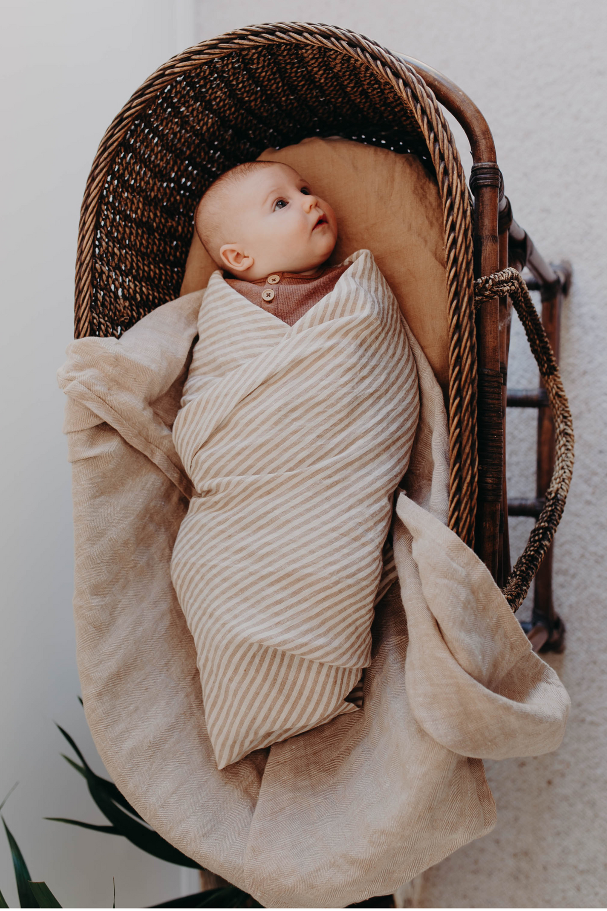Newborn snug in the Burleigh stripe linen swaddle, fabric spilling over the edge of a woven bassinet