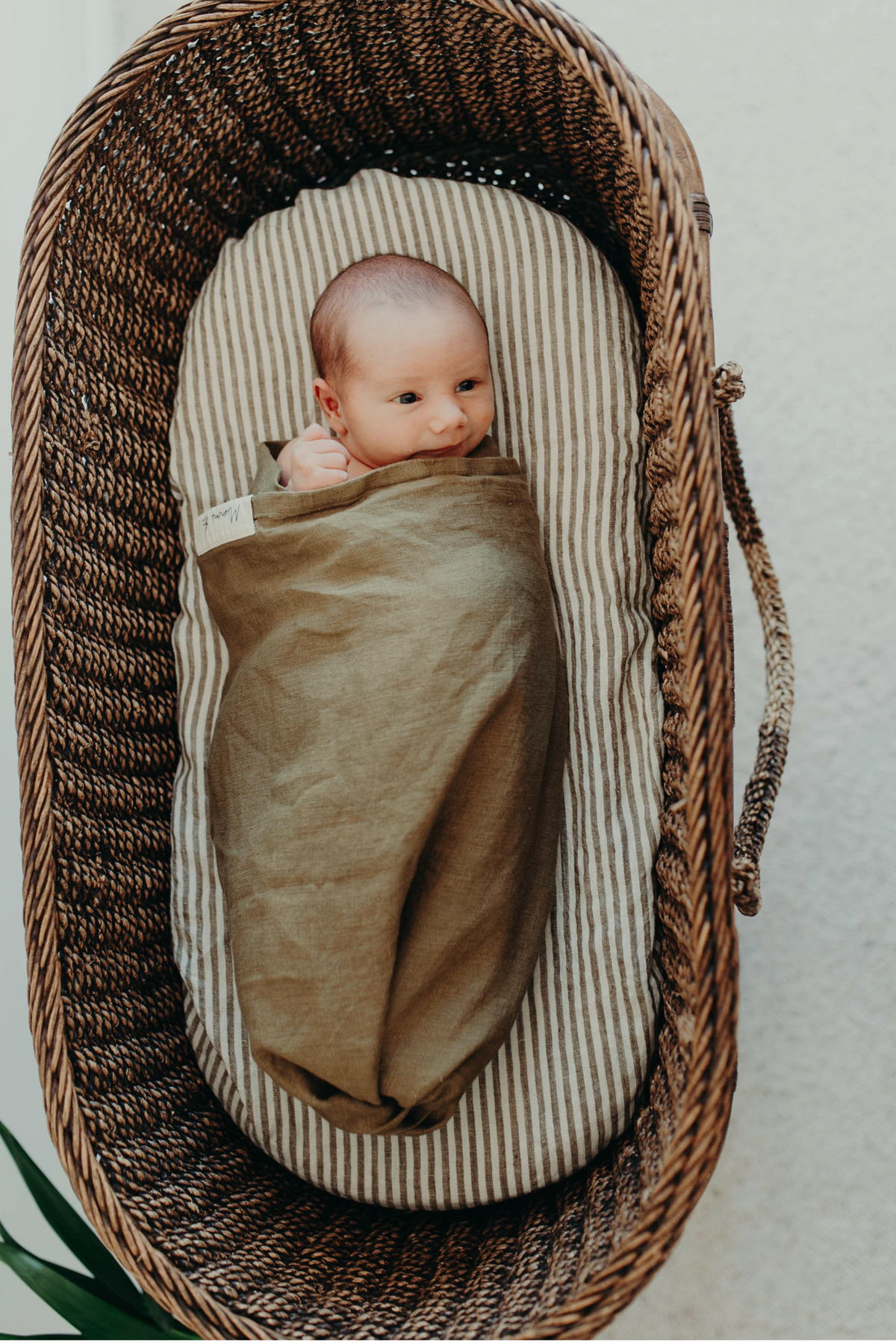 Newborn looking up from a woven bassinet, wrapped in the Willow linen swaddle with the woven label showing
