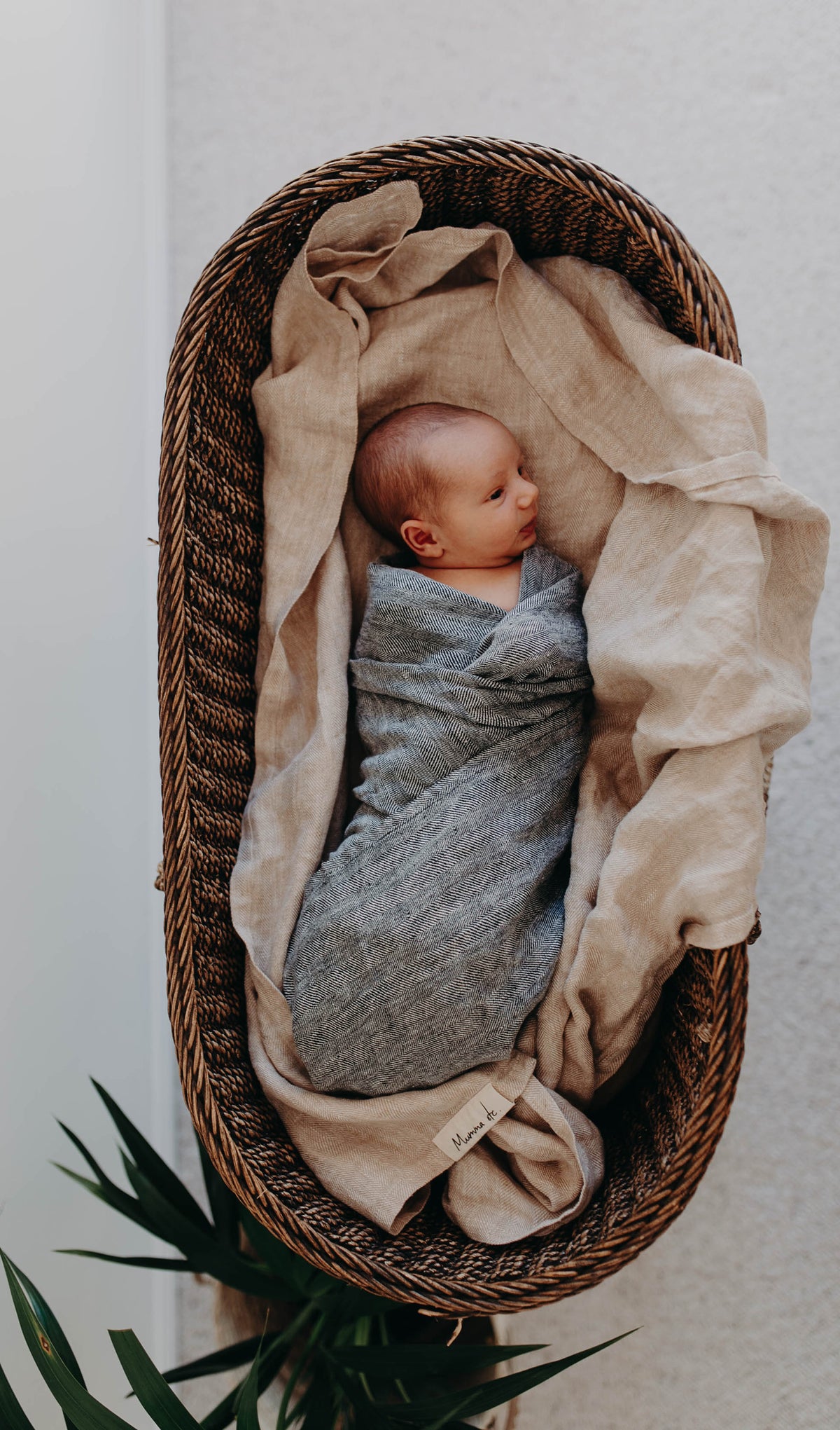 Newborn wrapped in the grey Aspen linen swaddle, lying on a sand linen blanket in a woven bassinet
