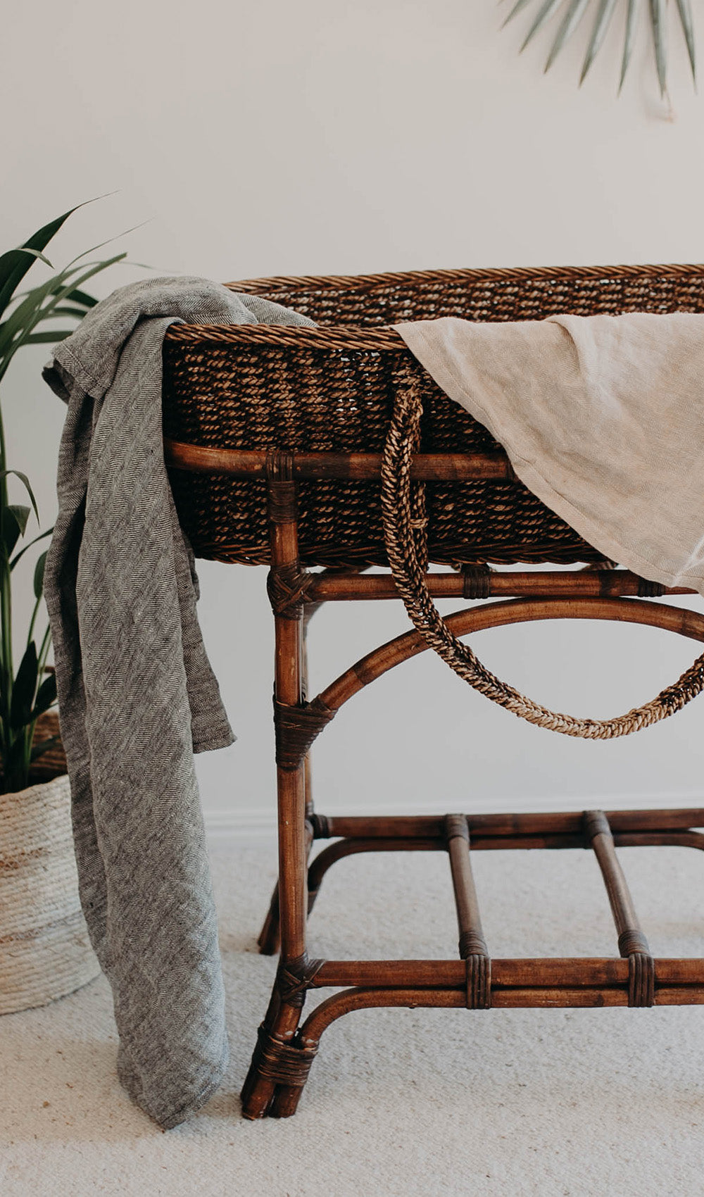 The Aspen linen swaddle draped over the side of a cane bassinet beside a potted palm