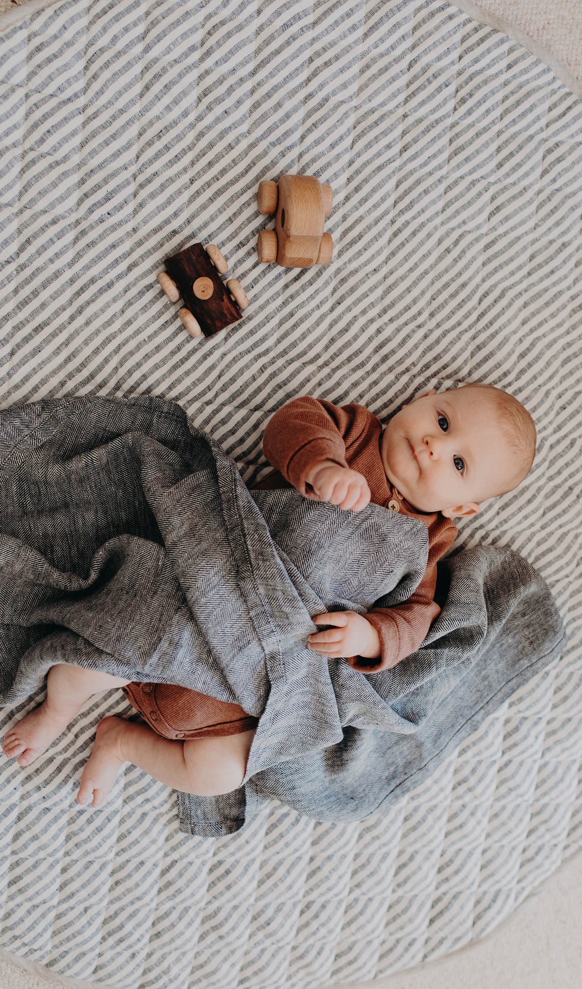 Baby in a rust romper lying on the unwrapped Aspen linen swaddle over a striped round rug, wooden toys beside