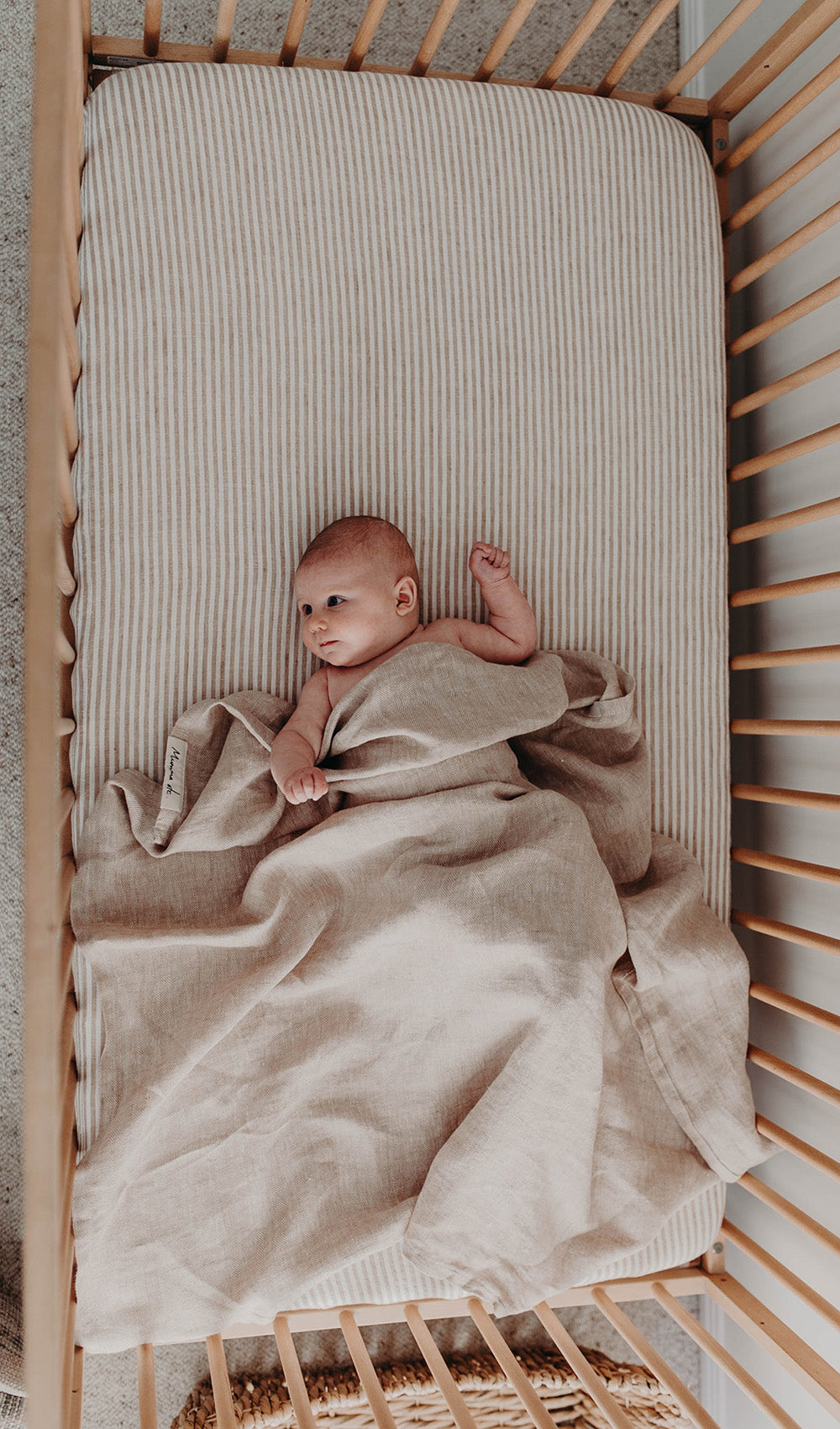 Newborn lying in a cot under the Meadow linen swaddle, one arm out and looking to the side