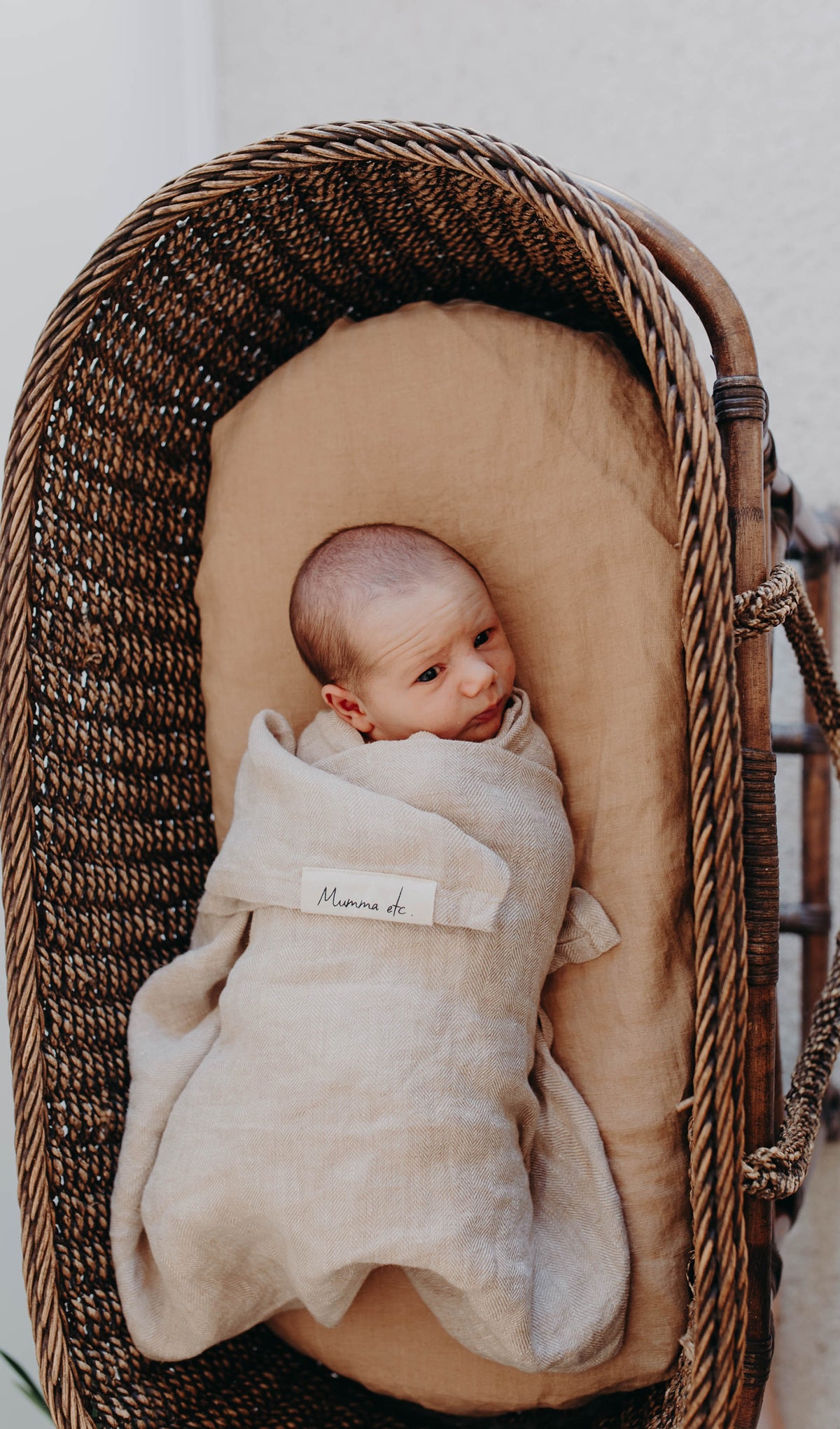 Newborn swaddled in the Meadow linen swaddle inside a cane bassinet, the woven label showing at the front