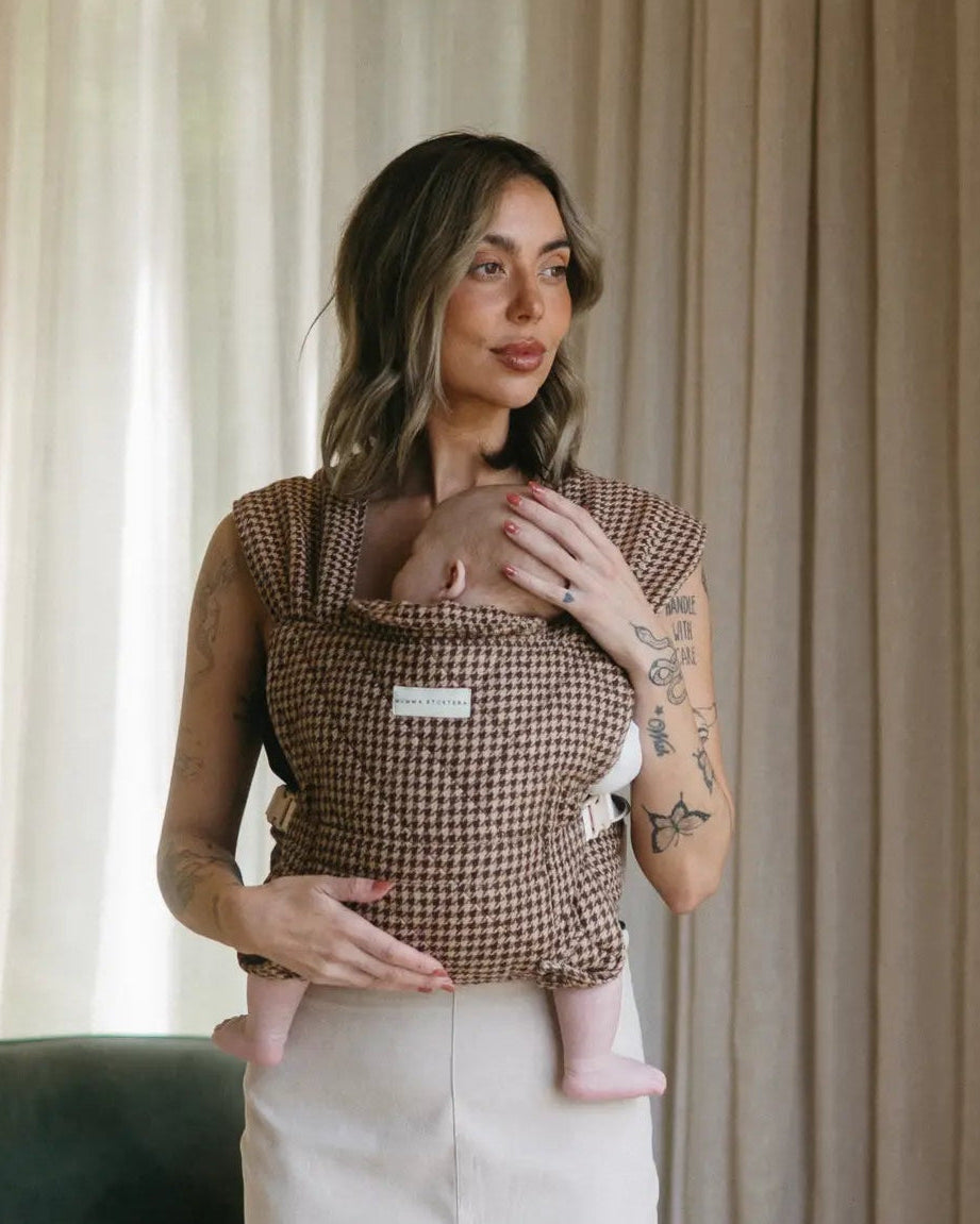 Mum by sheer curtains with her newborn carried facing inward in the Arista houndstooth Daily baby carrier, one hand on his head.