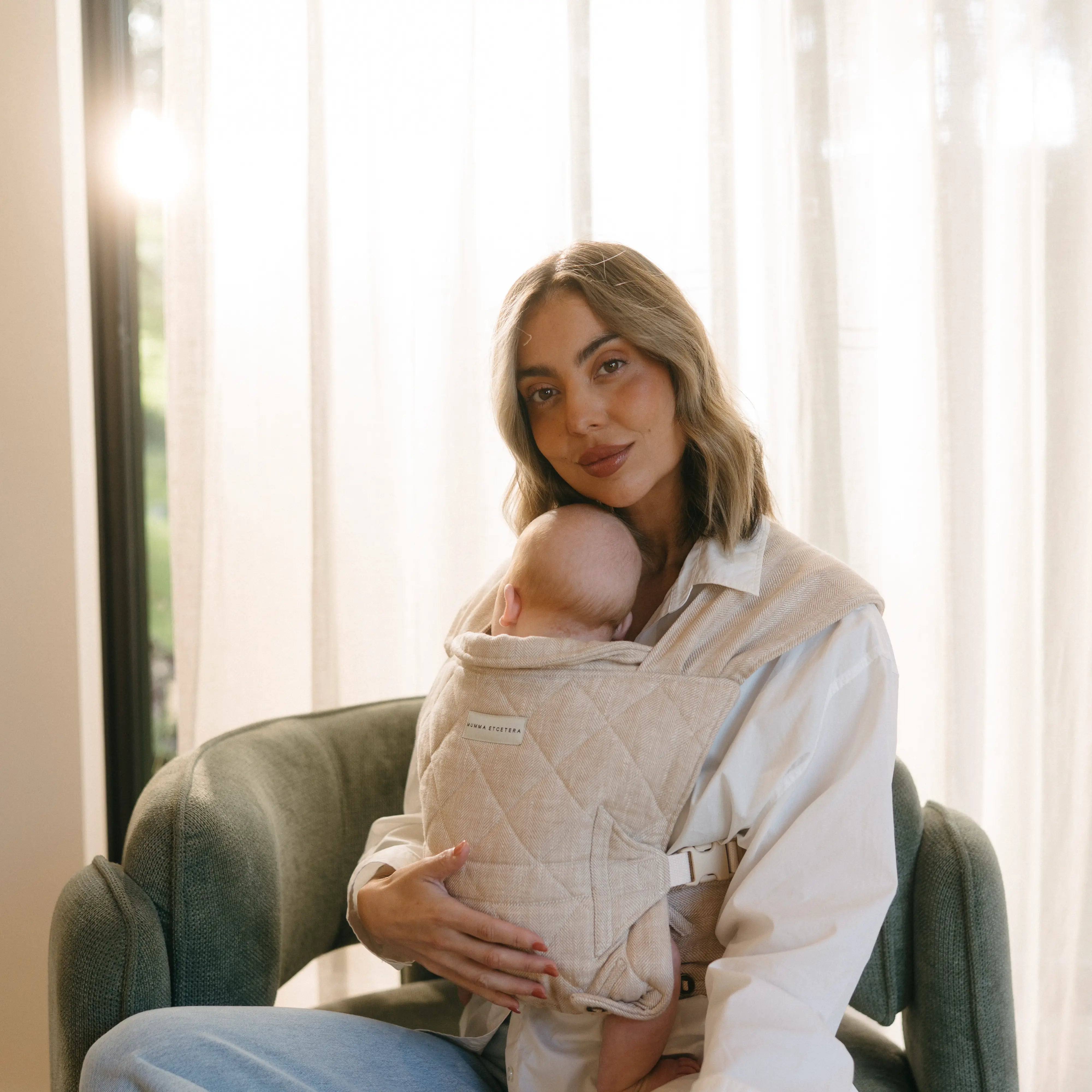 Mum tilts her head towards her sleeping newborn in the Meadow linen Daily baby carrier, sunlight through sheers
