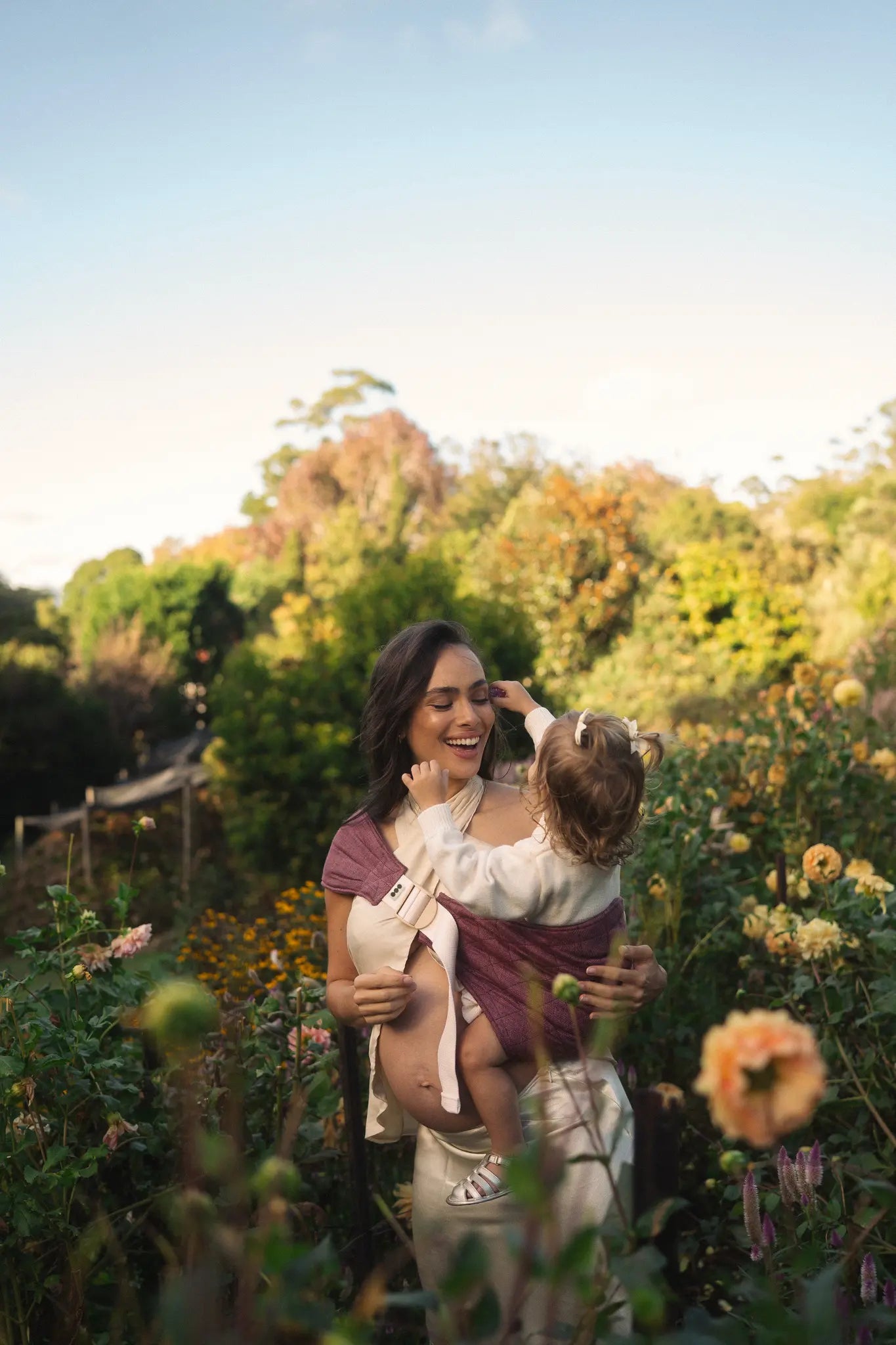 Toddler on her mum's hip in the Bloomè linen Hipsy toddler carrier, one hand supporting, among garden dahlias.