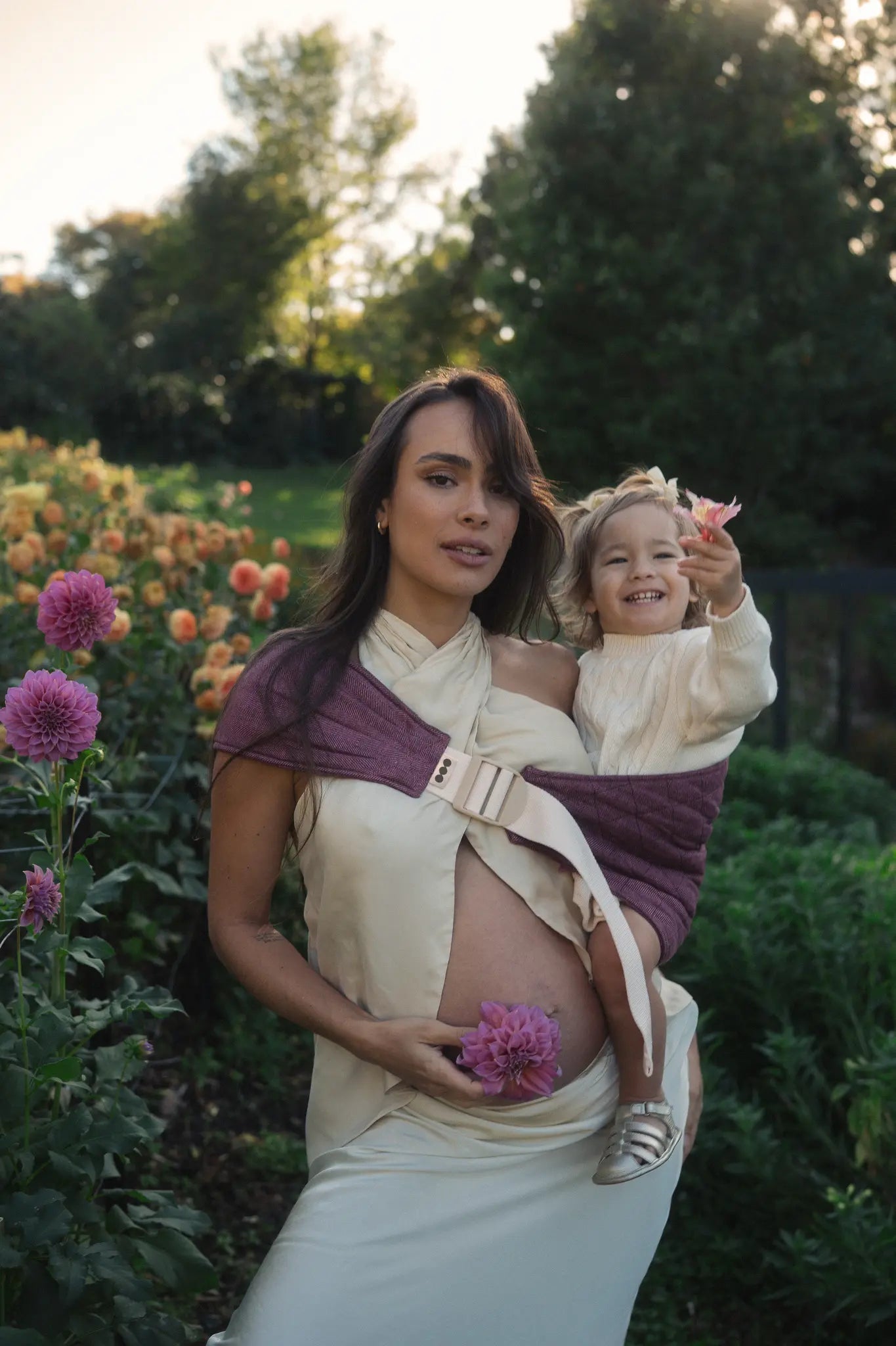 Mum carries her toddler on one hip in the Bloomè linen Hipsy toddler carrier beside the dahlia beds