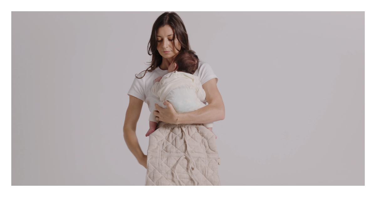 Mum lifting her newborn onto her chest above the linen Daily baby carrier panel, one hand supporting baby's head