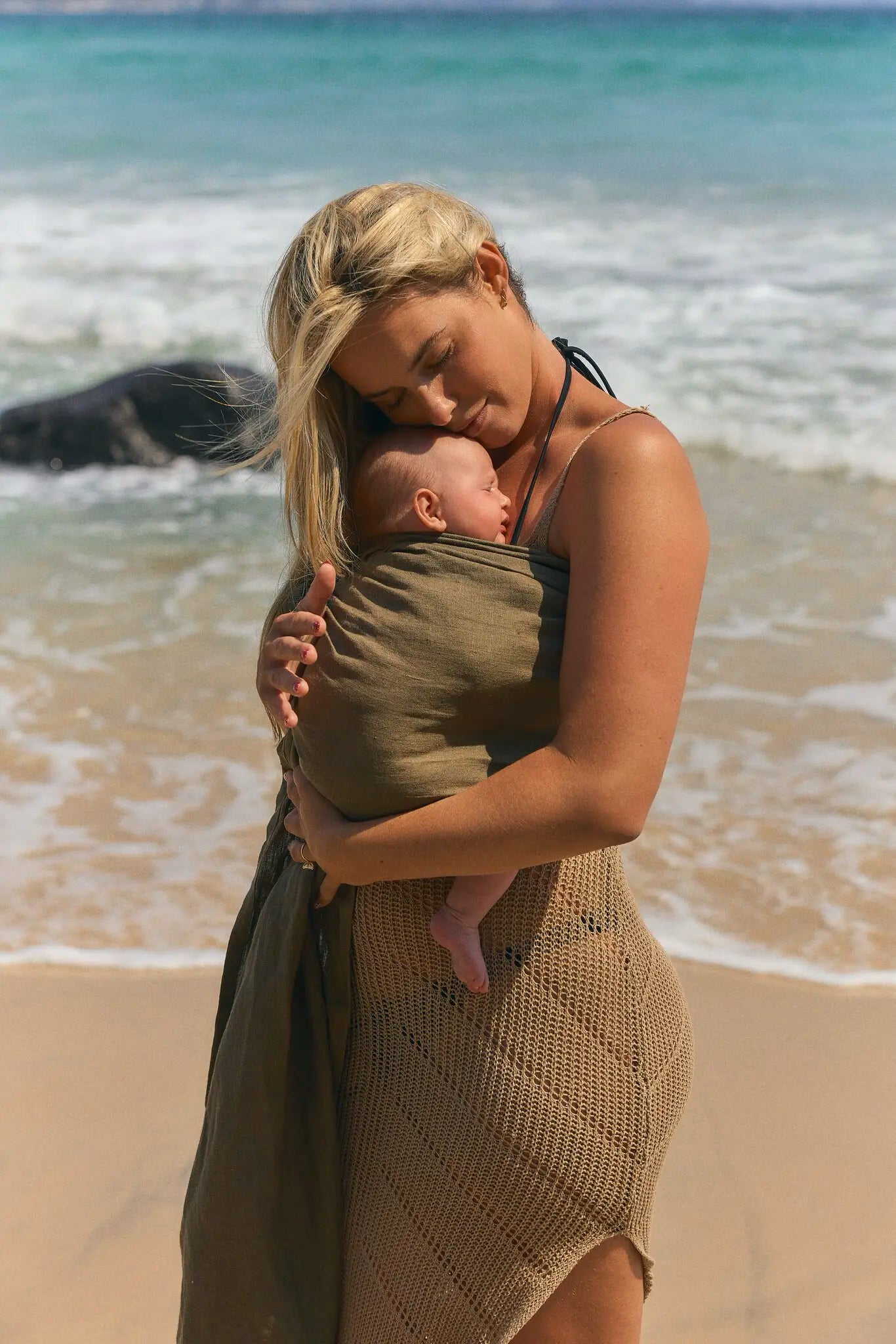 A mum leans in to kiss her newborn resting in the Aloe linen Sling baby carrier on a sunlit beach.