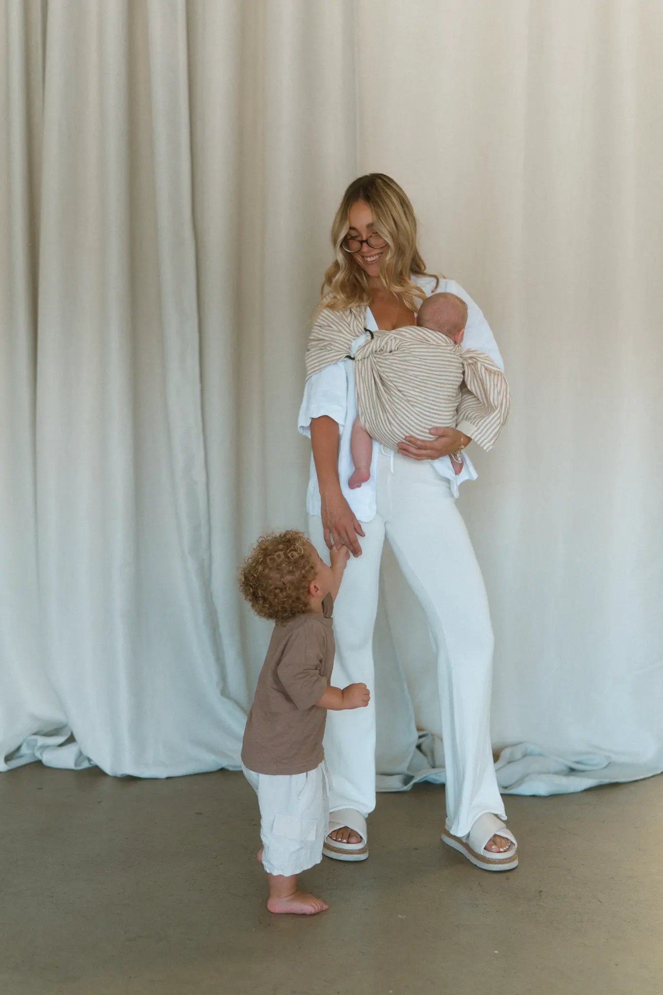 A mum holds her toddler's hand while her newborn rests in the Burleigh stripe Sling baby carrier.