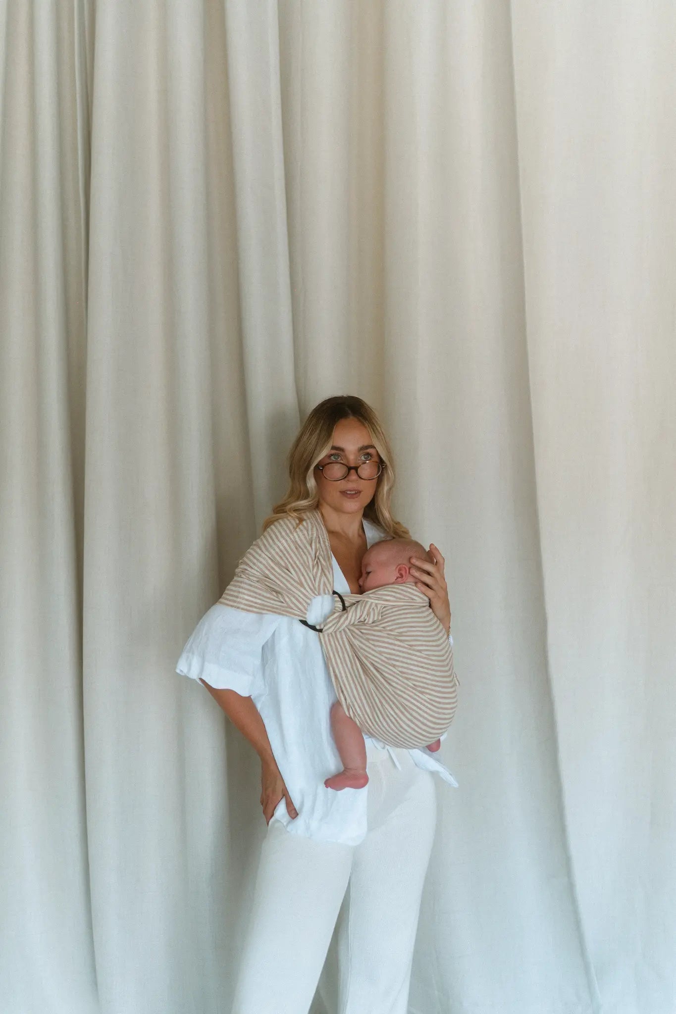 A mum steadies her newborn's head in the Burleigh stripe Sling baby carrier against a linen curtain.