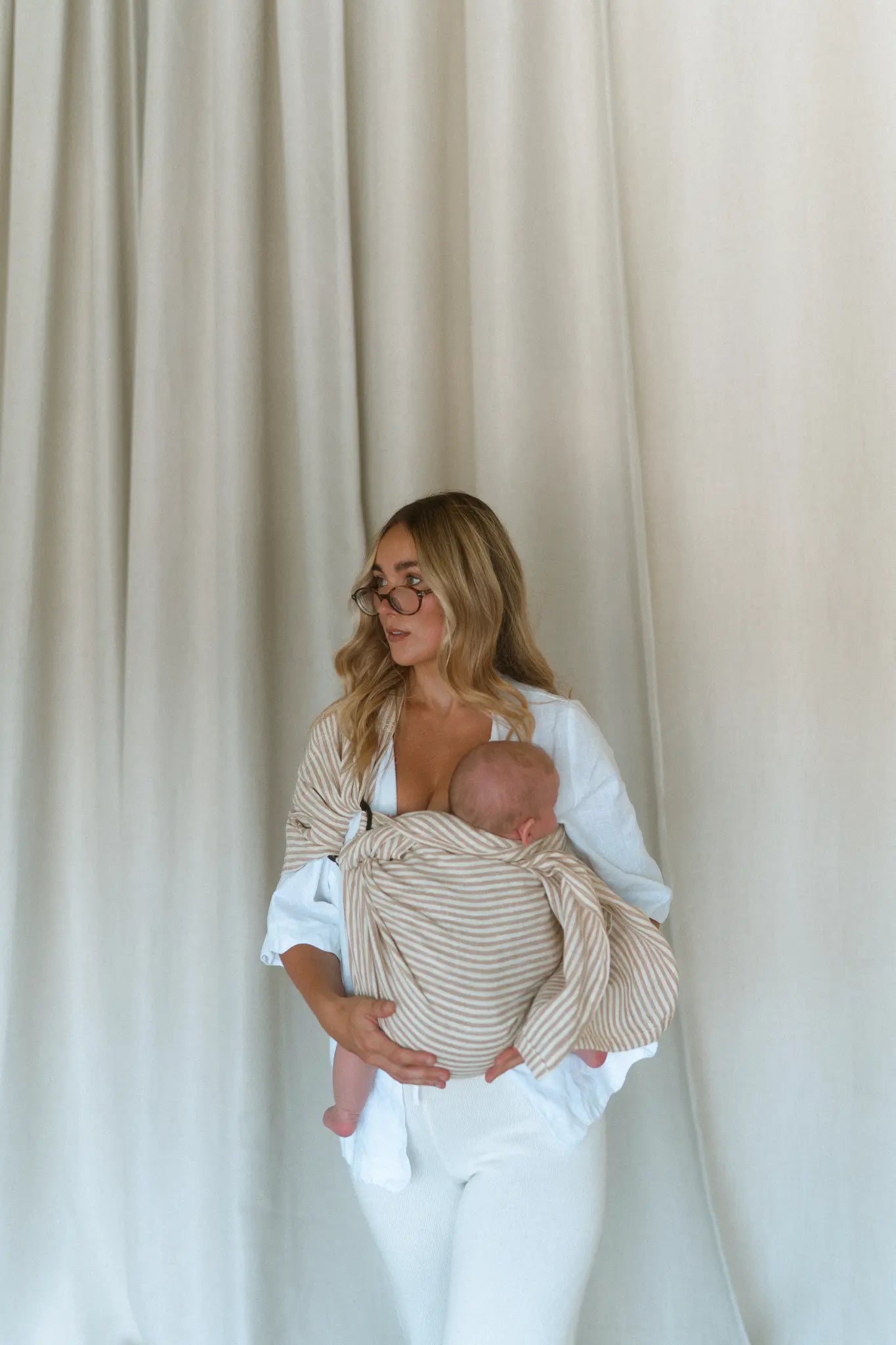 A mum looks off camera with her baby tucked into the Burleigh stripe Sling baby carrier, hand supporting.