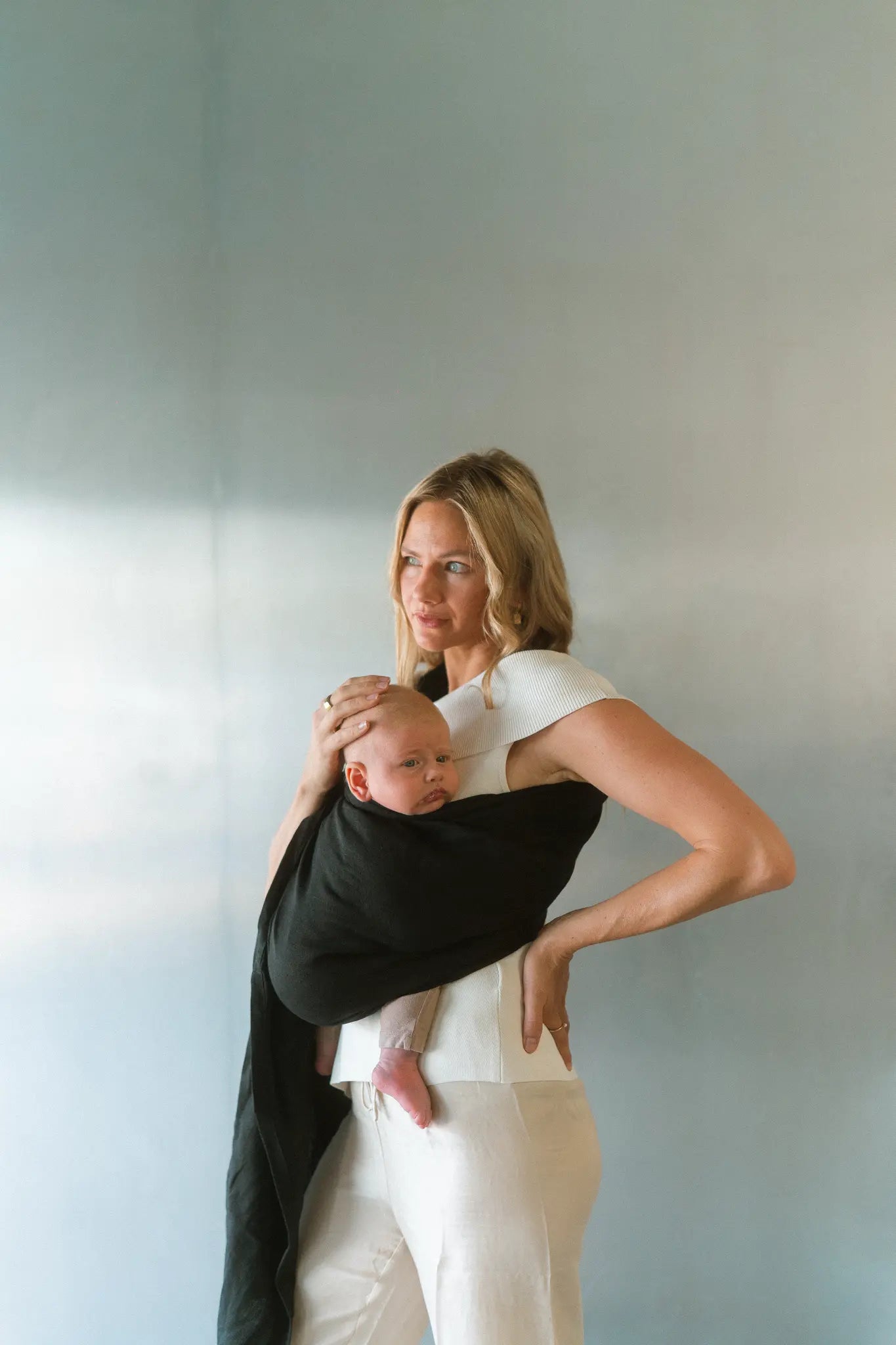 Hand resting on her baby's head, a mum wears the Tao linen Sling baby carrier against a pale wall.