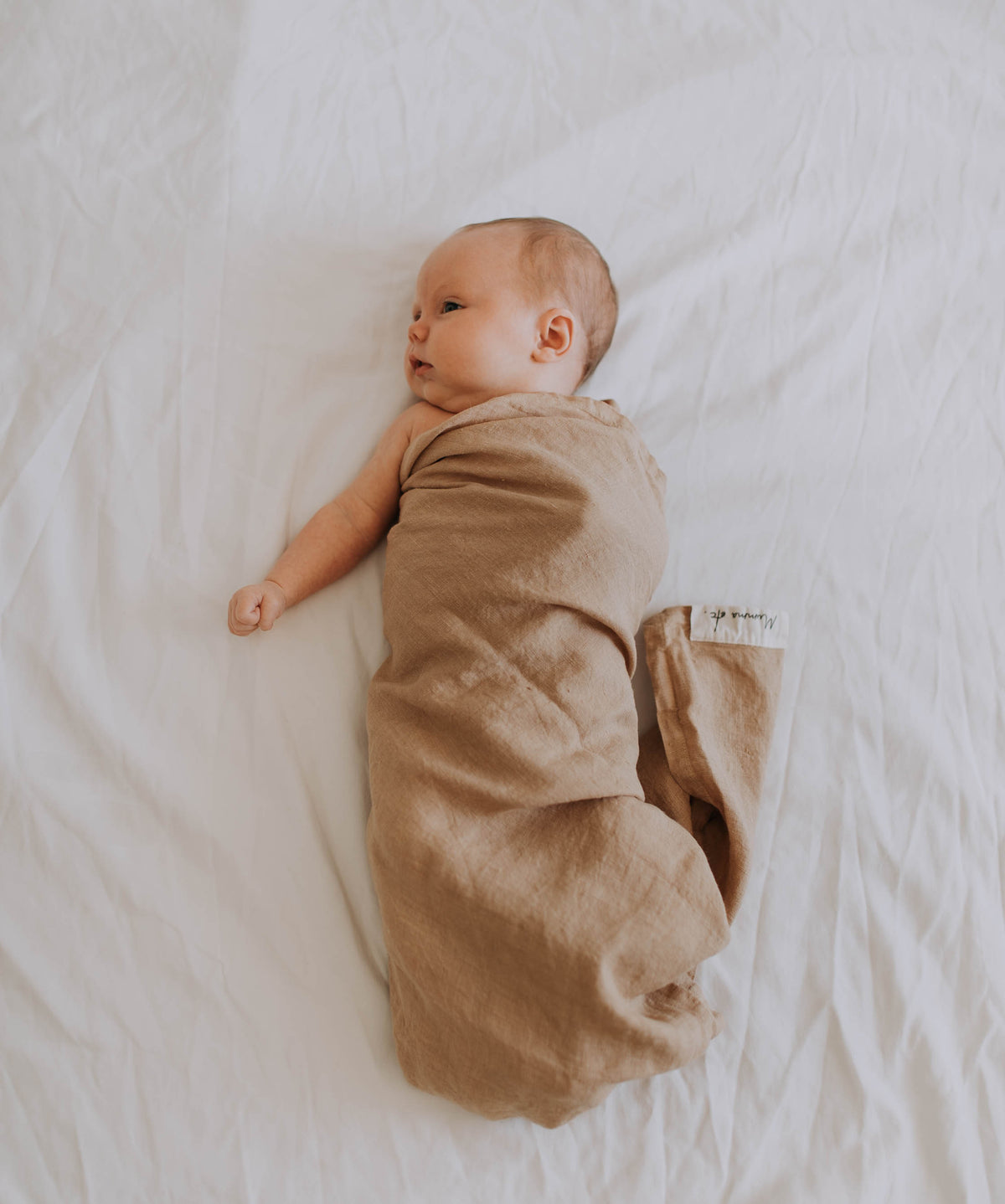 Newborn swaddled in a caramel linen swaddle, lying on a white sheet and looking up