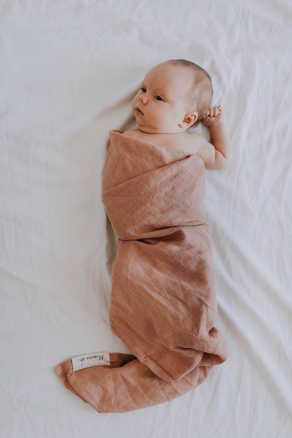 Newborn swaddled snugly in the Russet linen swaddle, dusty rose-rust against a white sheet