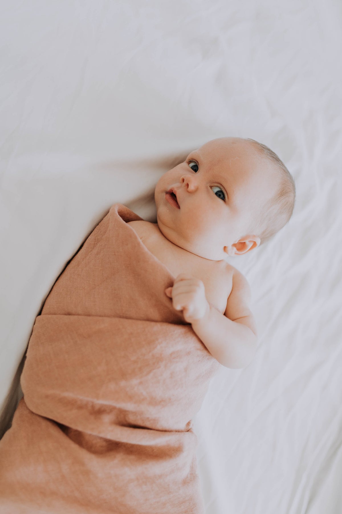 Newborn lying on a white sheet, loosely wrapped in the Russet linen swaddle and looking up
